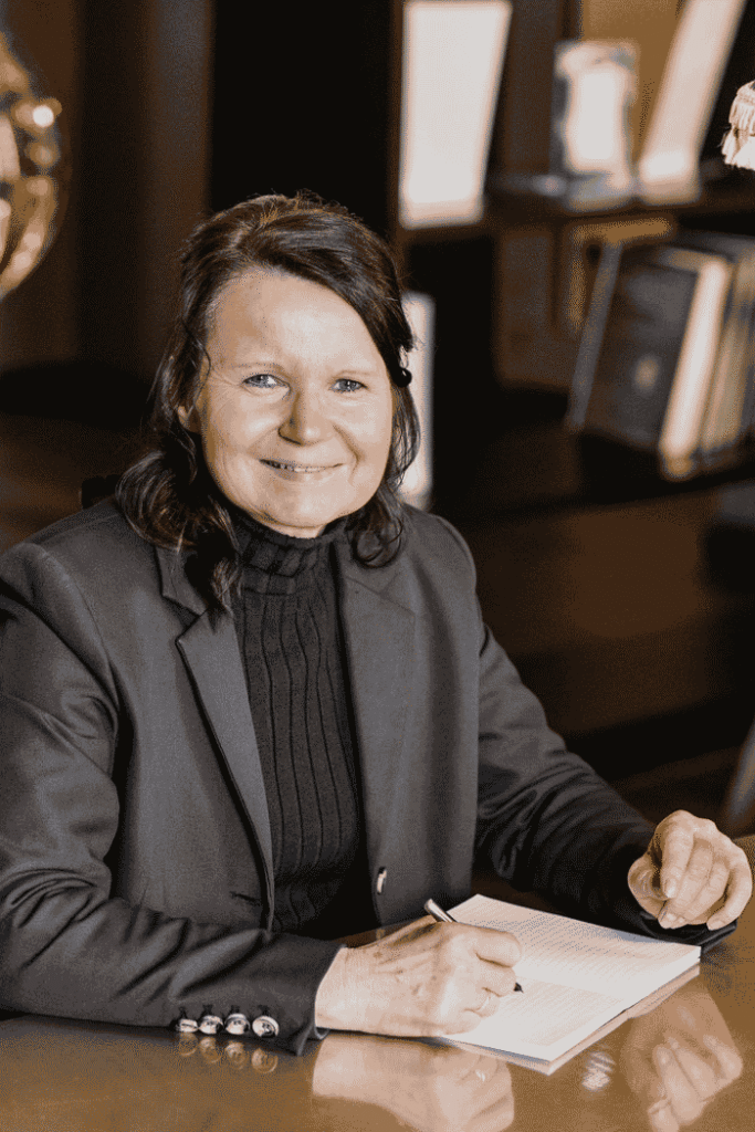 Sylvana Pollehn, Expertin für Service-Exzellenz, sitzt mit einem aufgeschlagenen Konzeptbuch in einer eleganten Hotel-Lobby und lächelt in die Kamera.